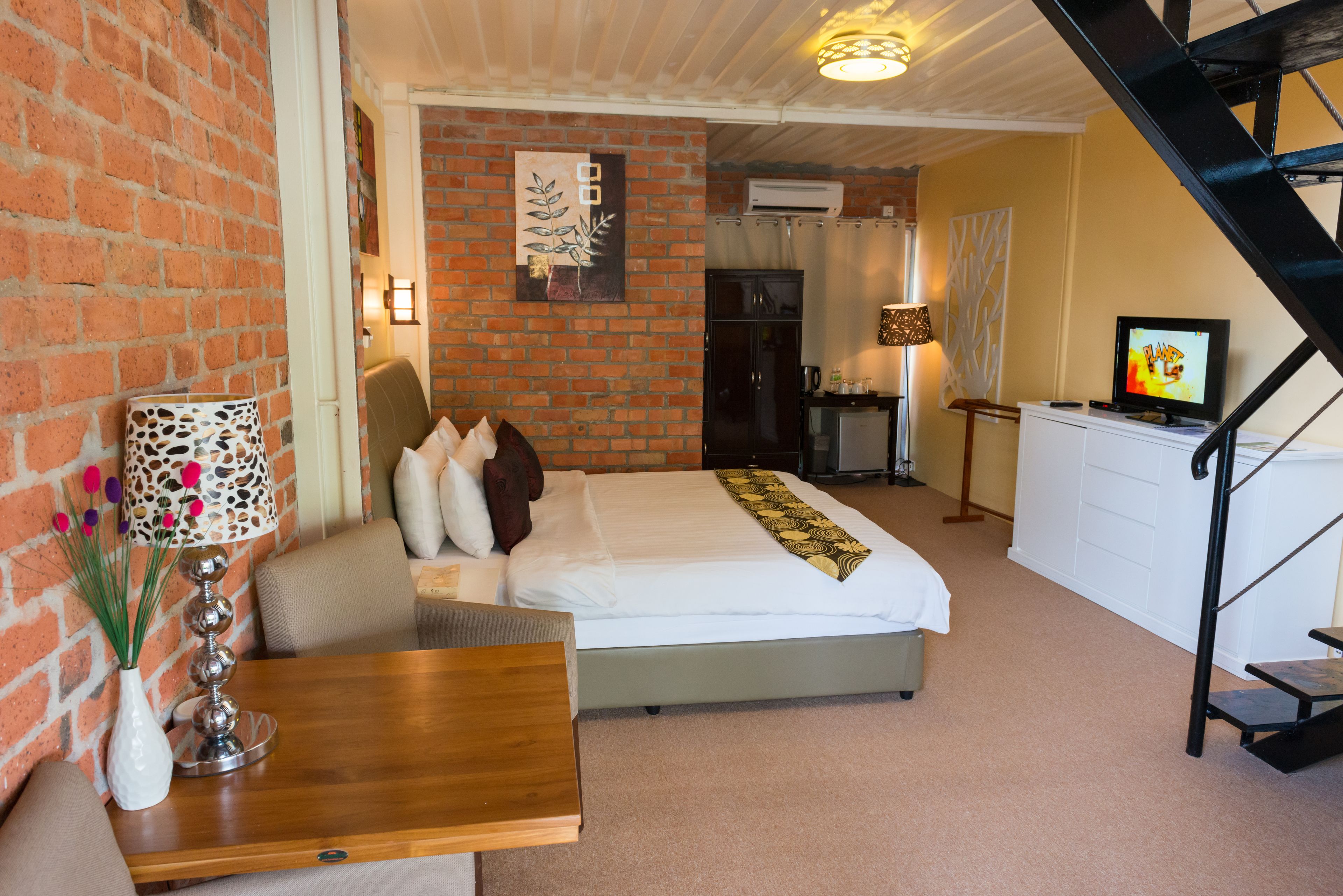 A bedroom with a brick wall, a bed, a desk, a lamp, and a TV.