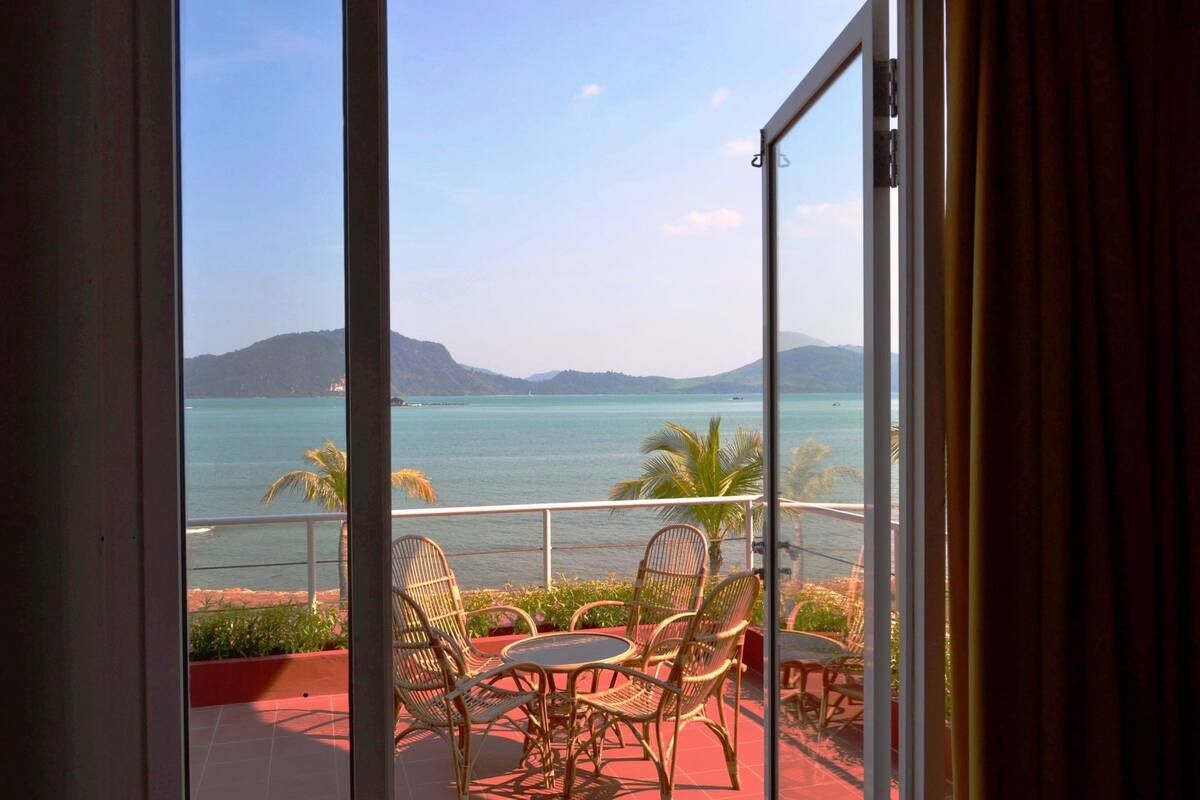 A balcony with wicker chairs and a table, overlooking a calm sea and distant mountains.