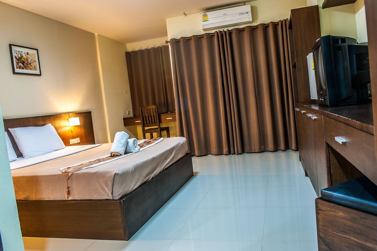 A hotel room with a bed, a desk, a chair, an air conditioning unit, and a television.