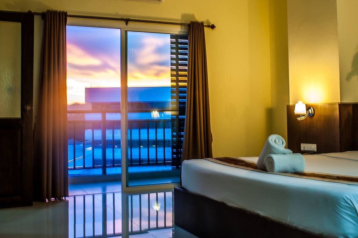 A hotel room with a bed, a nightstand, a lamp, an air conditioning unit, and a view of the ocean through a sliding door.