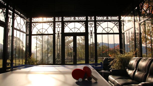 Uma mesa de ping pong com duas raquetes em um quarto com uma janela grande, um sofá de couro e uma mesa de tênis de mesa SpinShot.