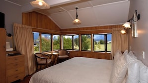 Quarto com cama grande, escrivaninha e cadeira, com janelas que dão para áreas verdes e montanhas.