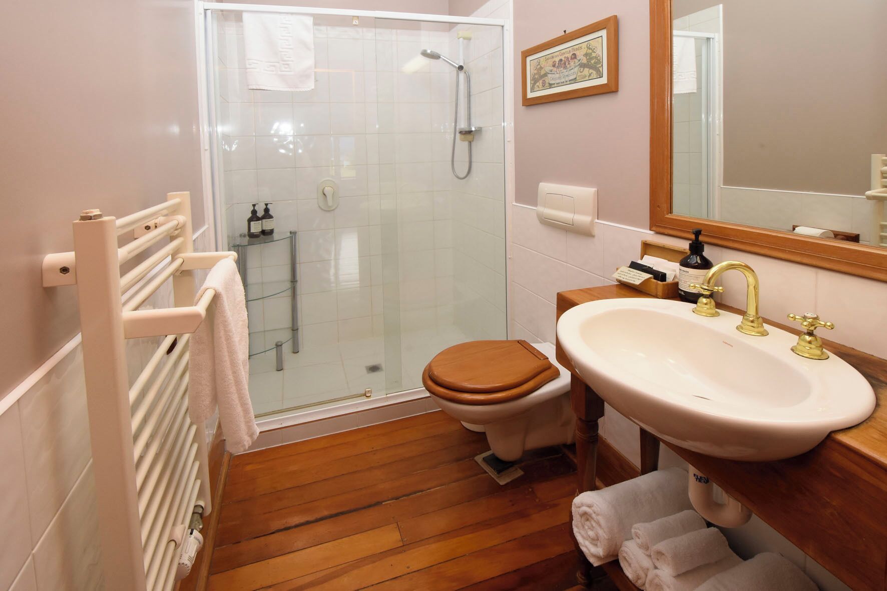 A bathroom with a glass shower enclosure, a wooden toilet, a white sink with gold faucets, and a wooden vanity.