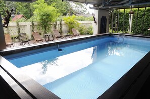Une piscine intérieure rectangulaire à l’eau d’un bleu limpide, entourée de chaises longues en bois et d’un espace abrité doté d’un toit en métal.