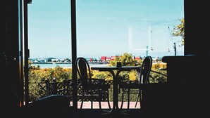 Une vue depuis une fenêtre, avec une table et des chaises, donnant sur une étendue d’eau et des bâtiments éloignés.