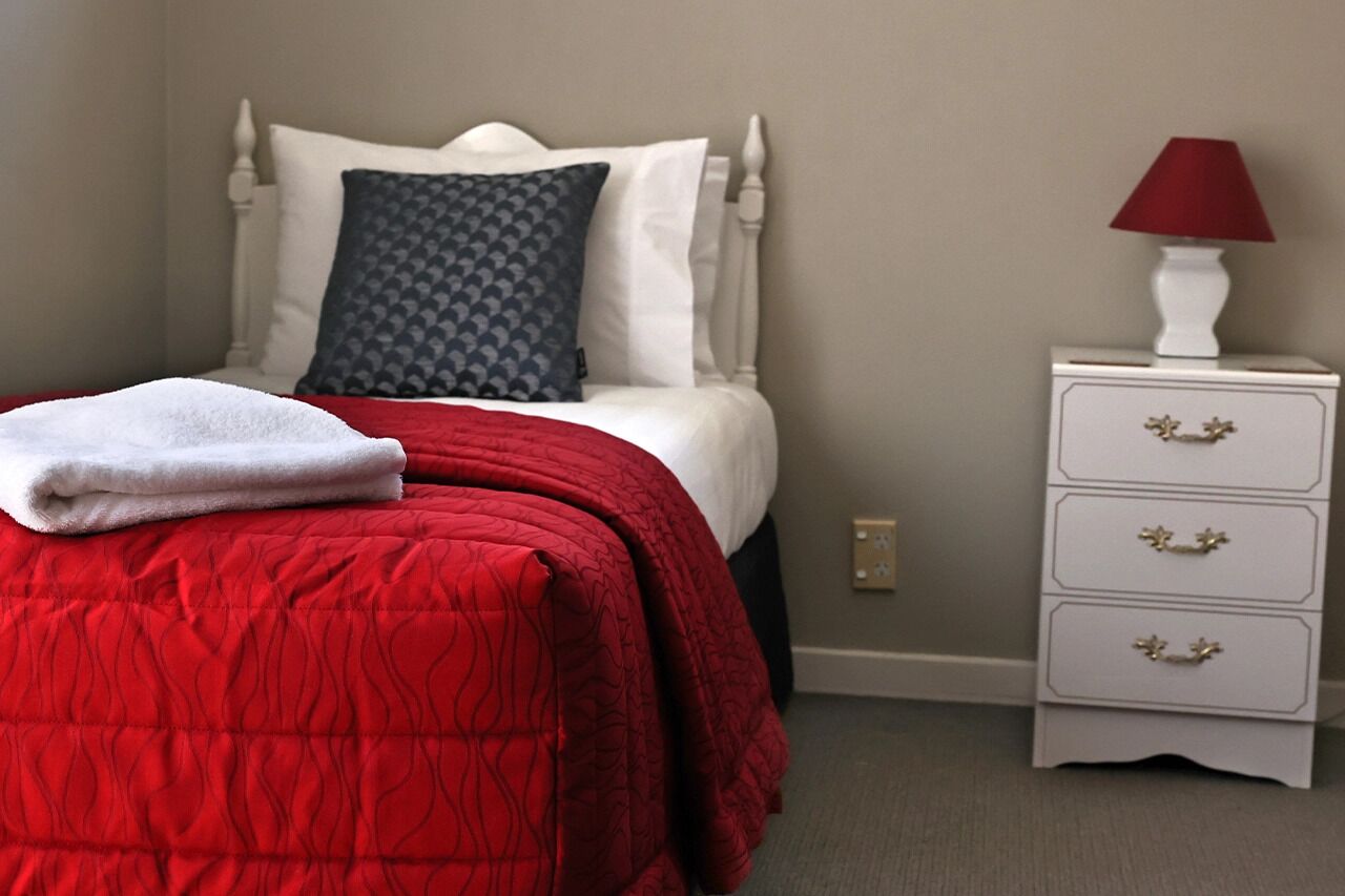 Un letto rifatto con cura, con una coperta rossa, biancheria bianca e un cuscino fantasia grigio. È presente un comodino bianco con una lampada rossa e tre cassetti con maniglie dorate.