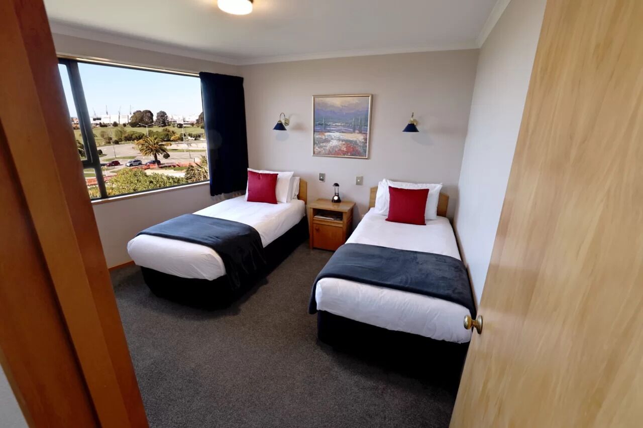 A hotel room with two beds, a nightstand, a painting, and a window with a view of the city.