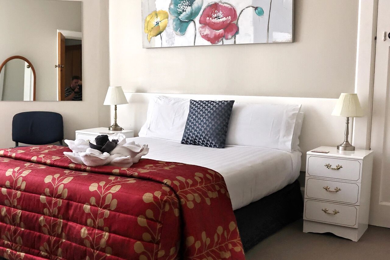A neatly made bed with a red floral bedspread, two bedside lamps, and a white nightstand with drawers.
