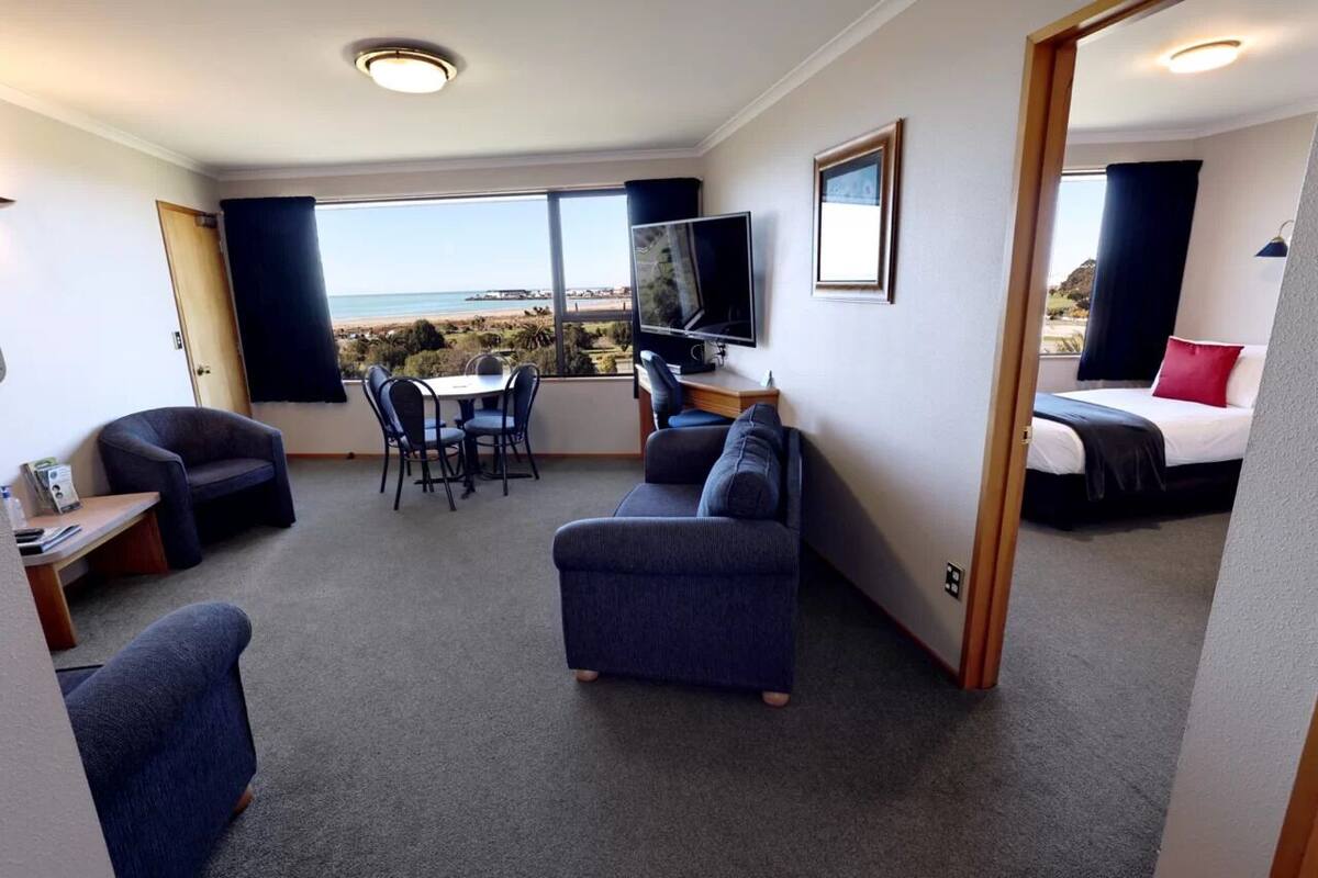 Ein Hotelzimmer mit einem großen Fenster und Blick auf den Strand, inklusive Fernseher, Bett, Sofa und Schreibtisch mit Stuhl.