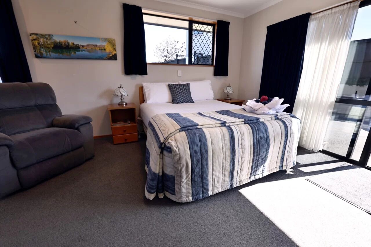 A hotel room with a bed, a chair, a nightstand, a window with curtains, and a painting on the wall.