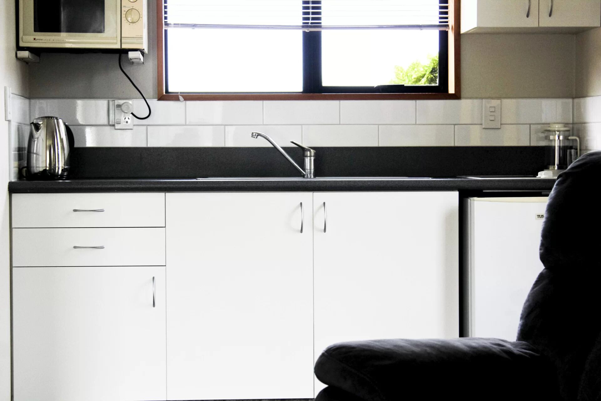 A compact kitchen with white cabinets, a black countertop, a kettle, and a window above the sink.