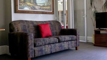 A living room with a patterned sofa, a red pillow, a television on a wooden stand, and a painting of a landscape on the wall.