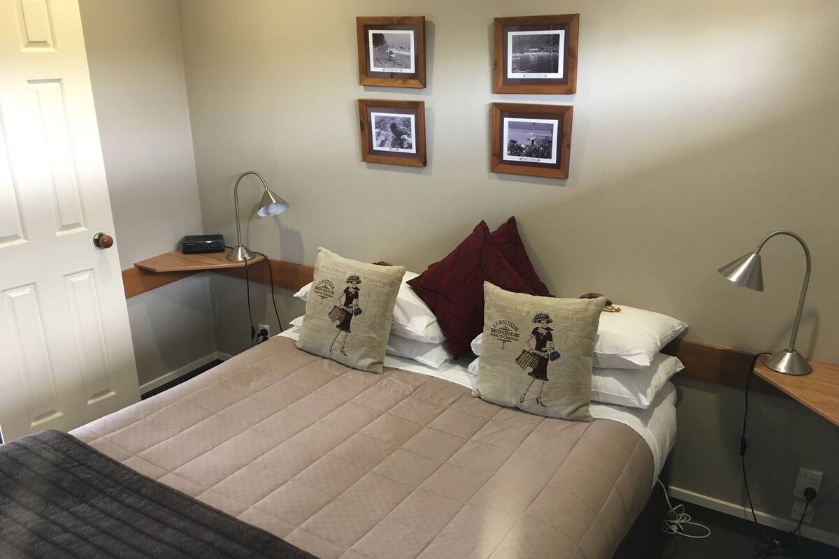 Una cama bien hecha con una colcha color beige y dos almohadones decorativos, una mesita de noche con lámpara y una pared con tres cuadros enmarcados.