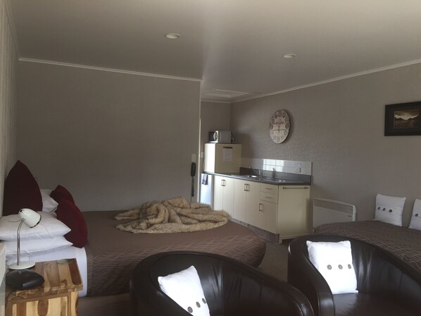 A hotel room with two beds, a kitchenette, a clock, and a small desk with a lamp.