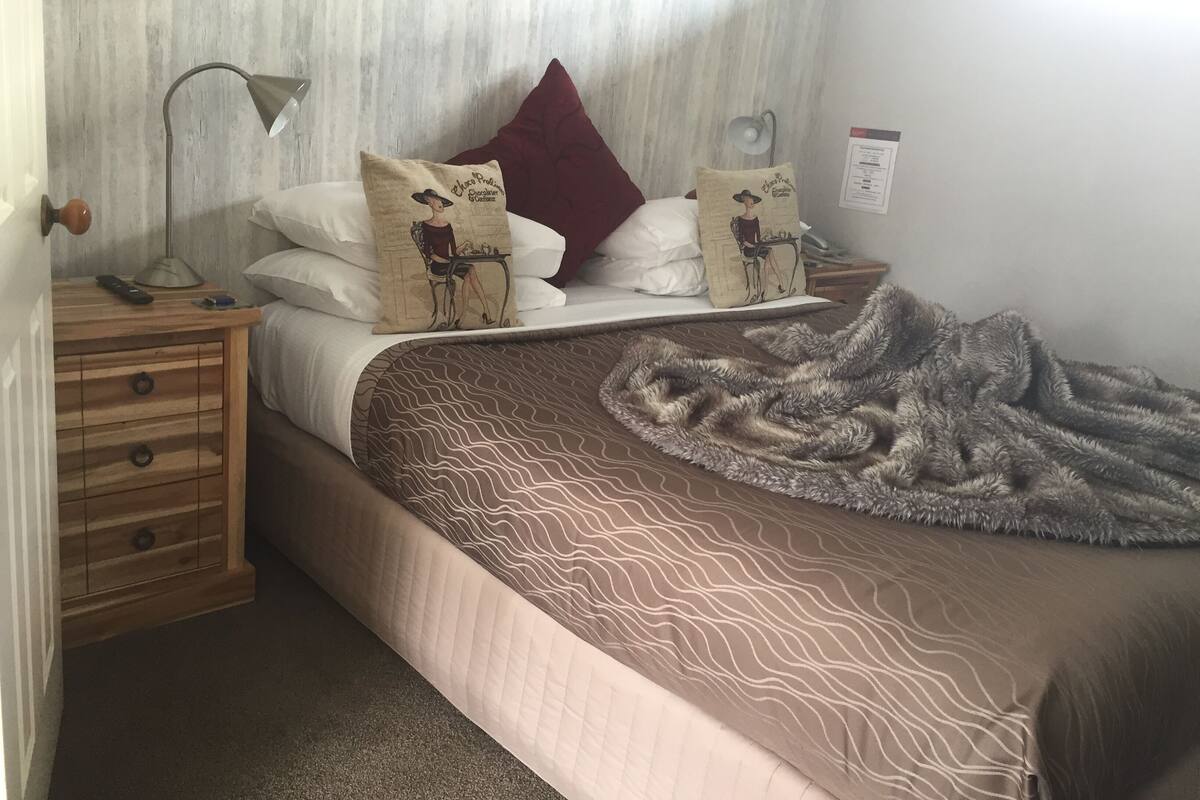 Una cama bien hecha con ropa de cama estampada, una mesita de noche de madera con una lámpara y un cojín rojo.
