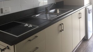 Una cocina con encimera negra, armarios blancos, un lavamanos de acero inoxidable y un microondas sobre un refrigerador.