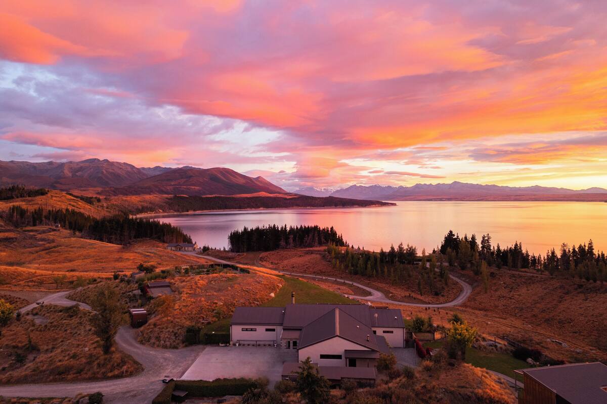 A house with a driveway, a small lake, and mountains in the background.