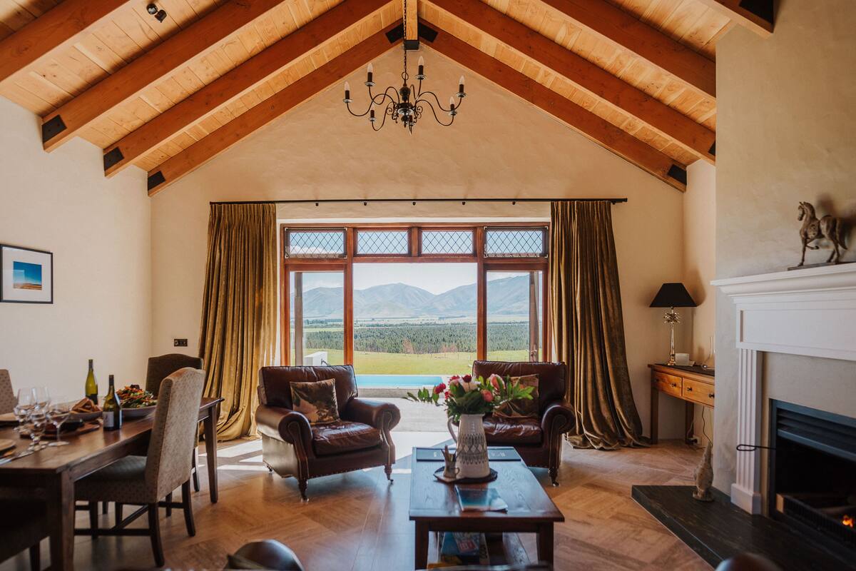 A spacious living room with a high wooden ceiling, a fireplace, and a large window offering a mountain view.