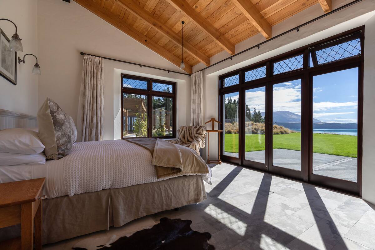 A bedroom with a large bed, a wooden ceiling, and a view of a green landscape through large windows.