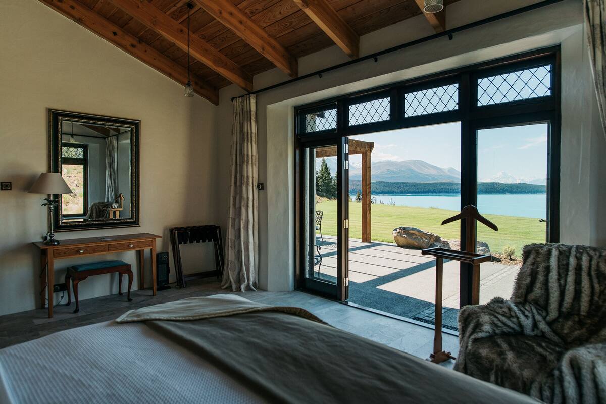 A bedroom with a large bed, a desk with a chair, a mirror, and a view of a lake and mountains through a glass door.