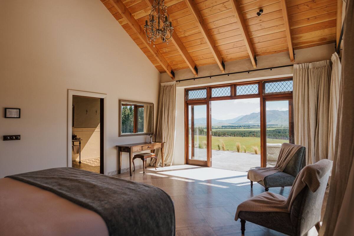 A spacious bedroom with a large bed, a desk, and a chair, with a view of the outdoors through large windows.