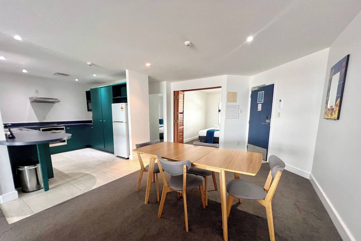 A modern kitchen with a built-in refrigerator, a dining area with a wooden table and four chairs, and a separate bedroom visible through an open door.
