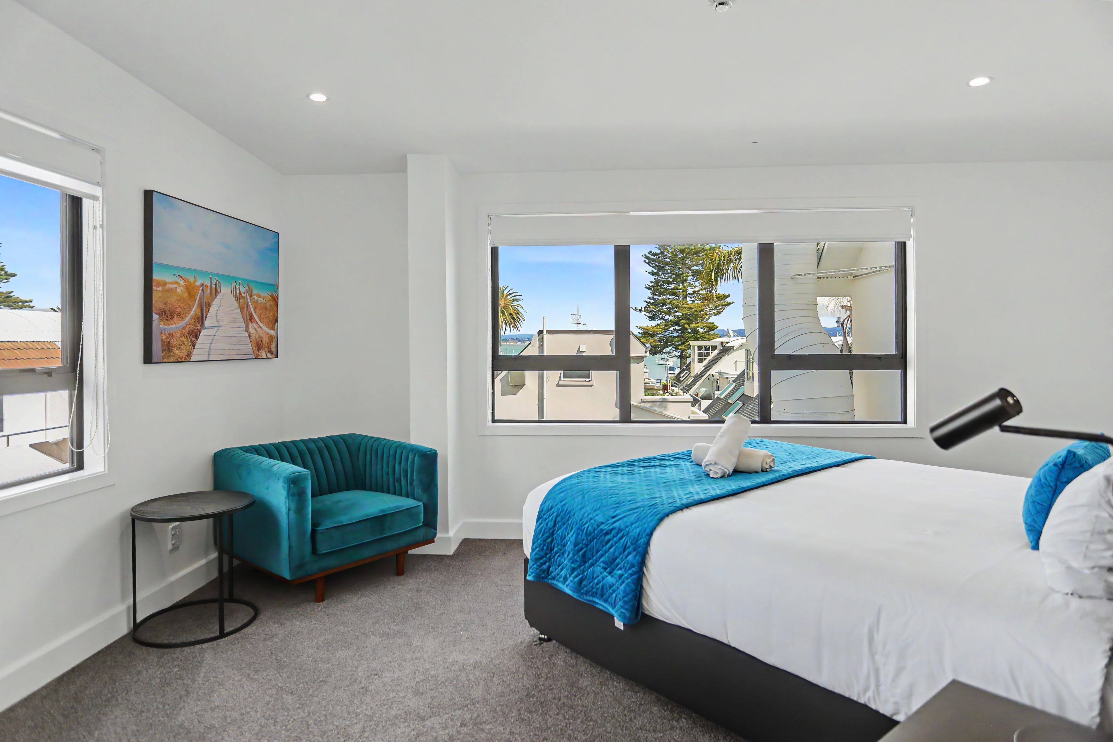 A hotel room with a bed, a blue sofa, a small table, a painting, and a window with a view of buildings and trees.
