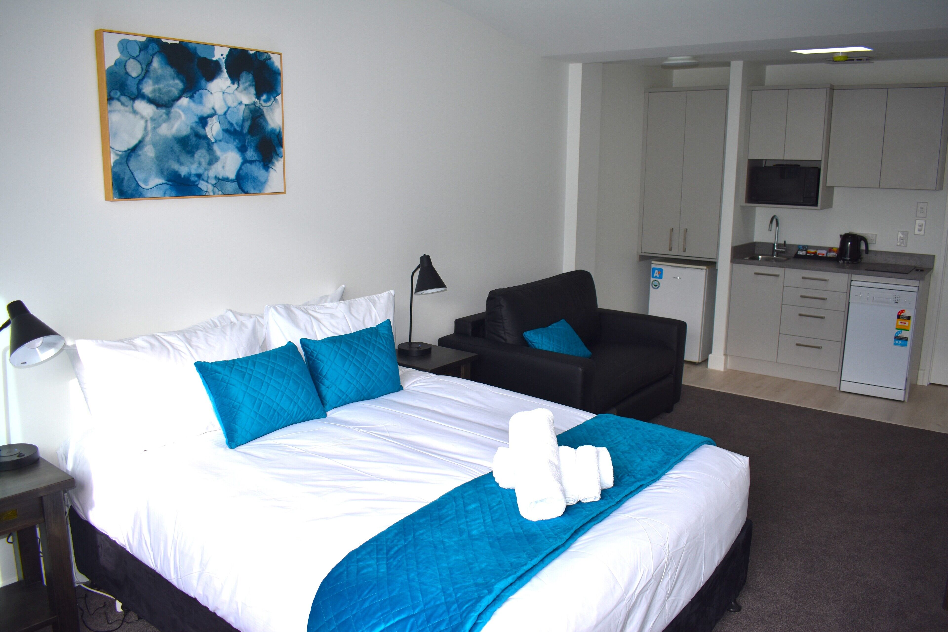 Quarto de hotel com cama, poltrona, escrivaninha, luminária e cozinha compacta.