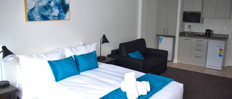 Quarto de hotel com cama, poltrona, escrivaninha, luminária e cozinha compacta.