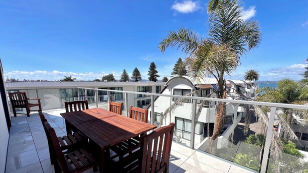 A balcony with a wooden table and chairs, overlooking a residential area with palm trees and a view of the sea. - 6 on Adams  (Tauranga)