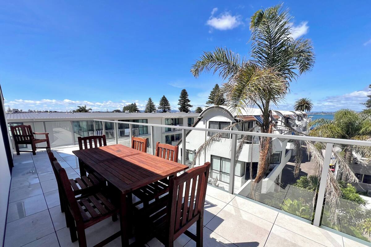 A balcony with a wooden table and chairs, overlooking a residential area with palm trees and a view of the sea.