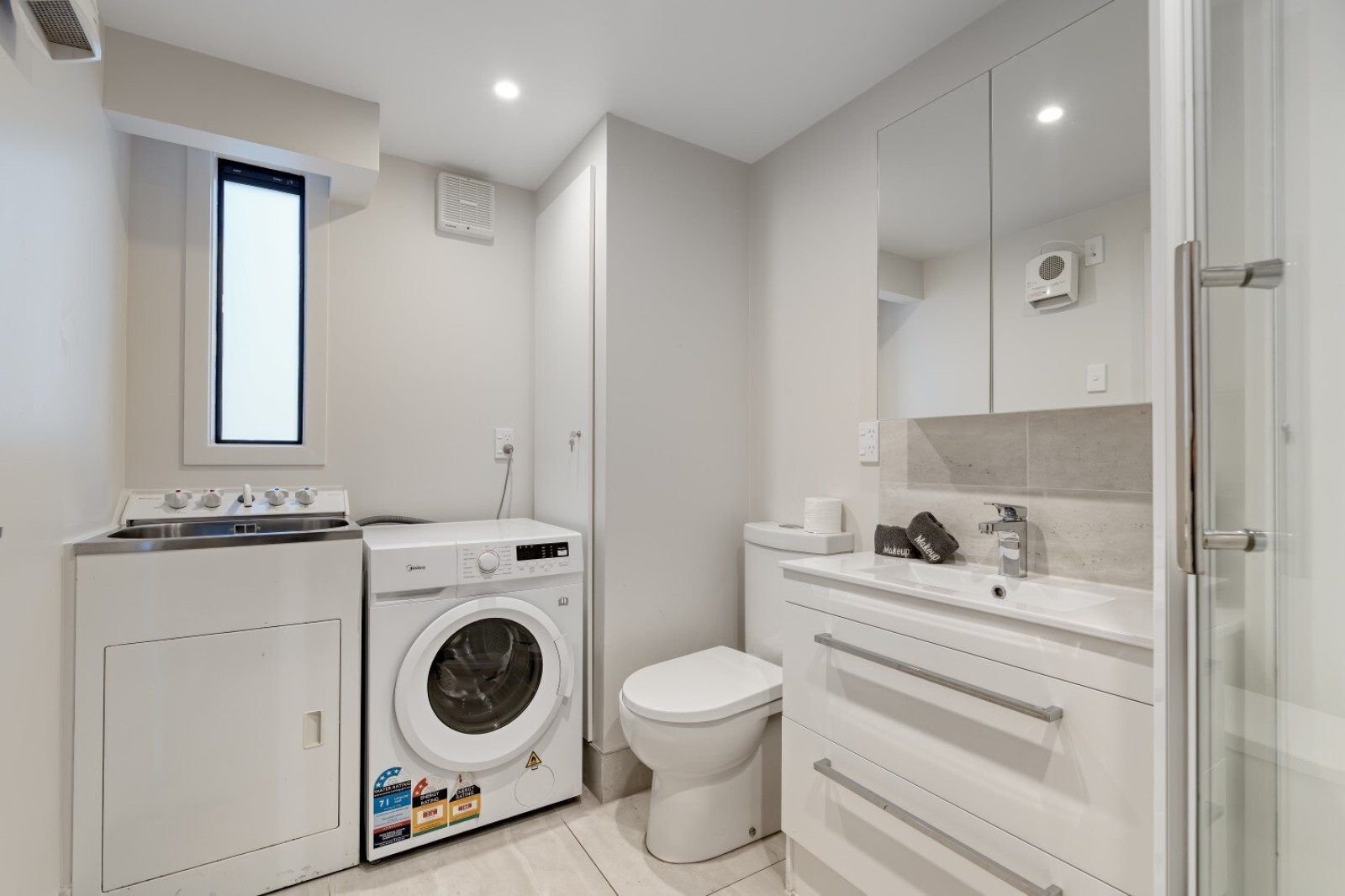 A modern bathroom with a washing machine, toilet, and a sink with a mirror above.