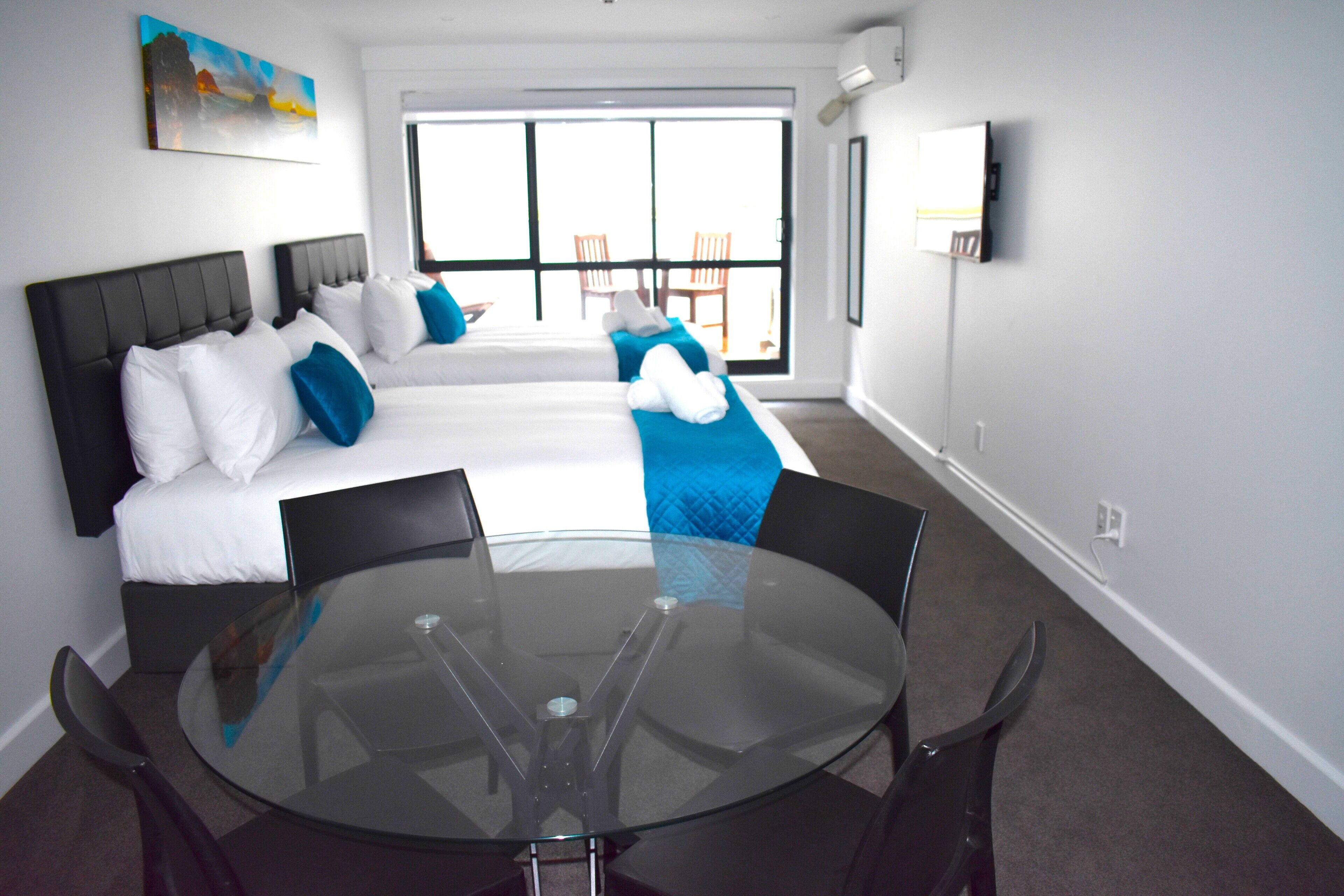 A hotel room with a large bed, a glass table, and a view of the outdoors.