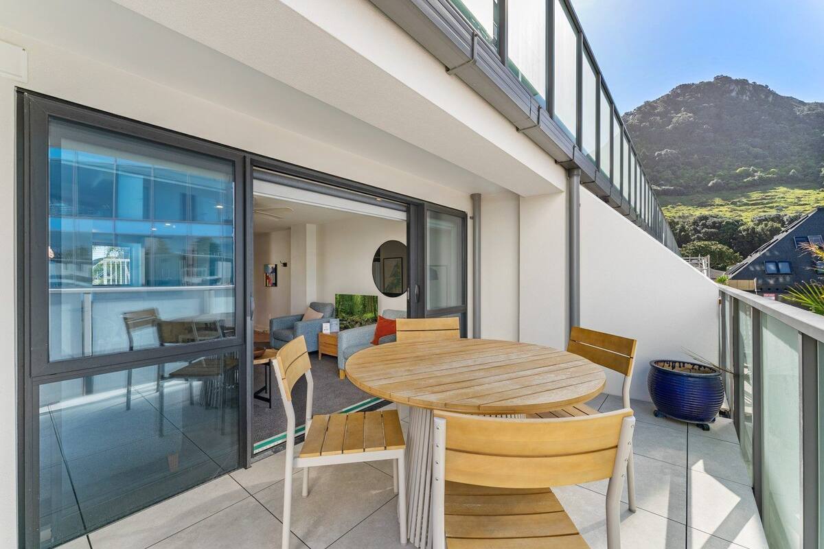 A modern outdoor balcony with a wooden table and chairs, a large blue pot, and a view of a mountain.