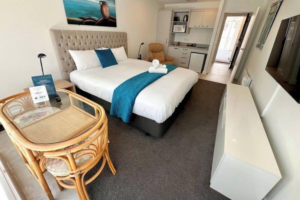 A hotel room with a bed, a desk, a chair, a small kitchenette, and a painting on the wall.