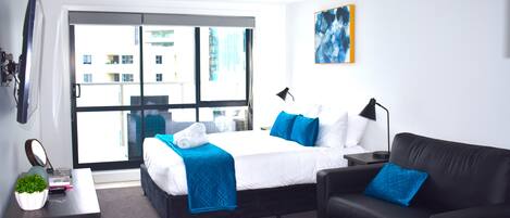 Una habitación de hotel moderna con una cama, un sofá de cuero negro, una pequeña mesa con una planta y un ventanal con vistas a la ciudad.