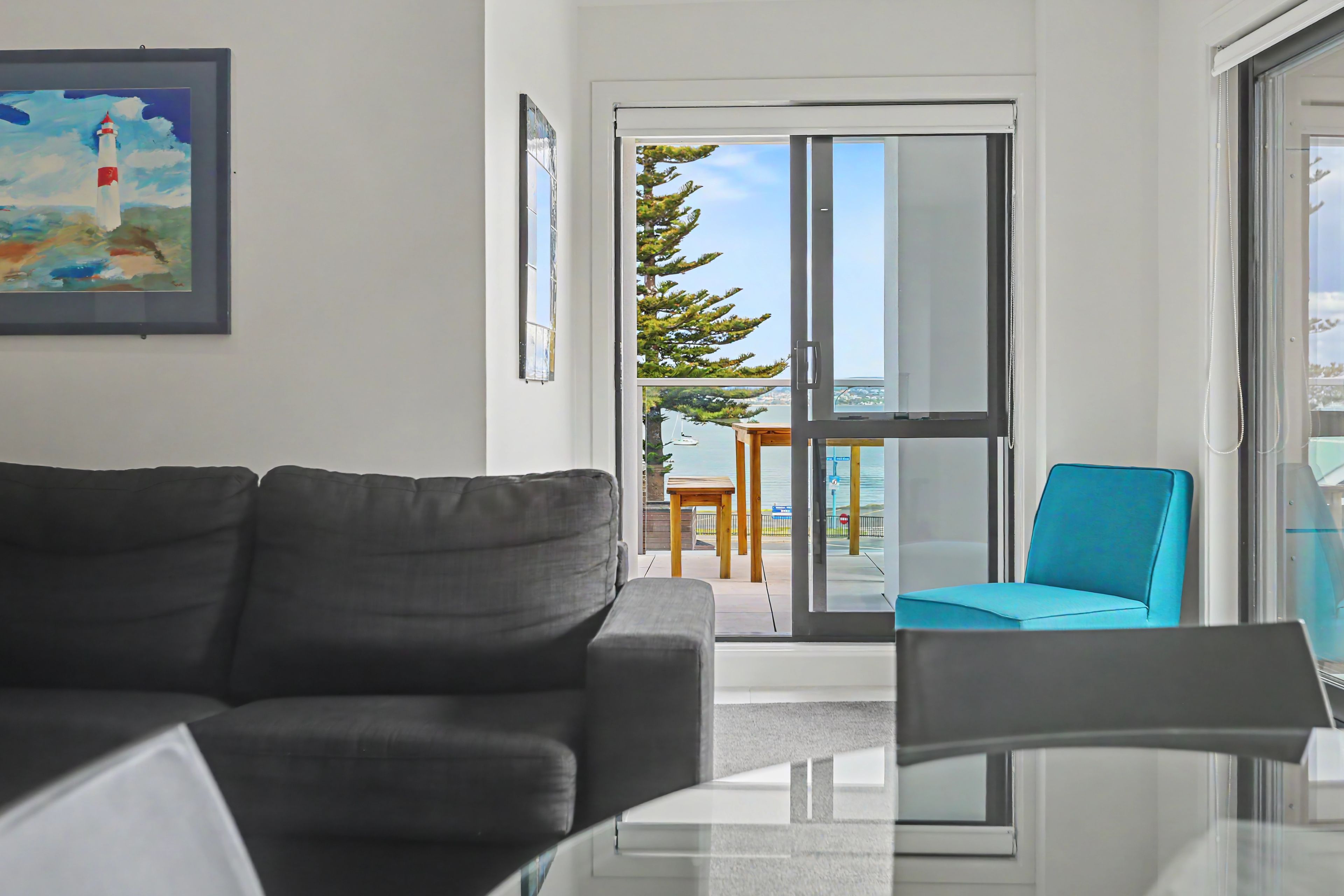 A modern living room with a dark-colored sofa, a glass coffee table, and a teal chair. A painting of a lighthouse is on the wall.