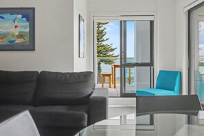Una sala moderna con un sofá oscuro, una mesa de centro de vidrio y una silla color turquesa. En la pared, un cuadro de un faro.