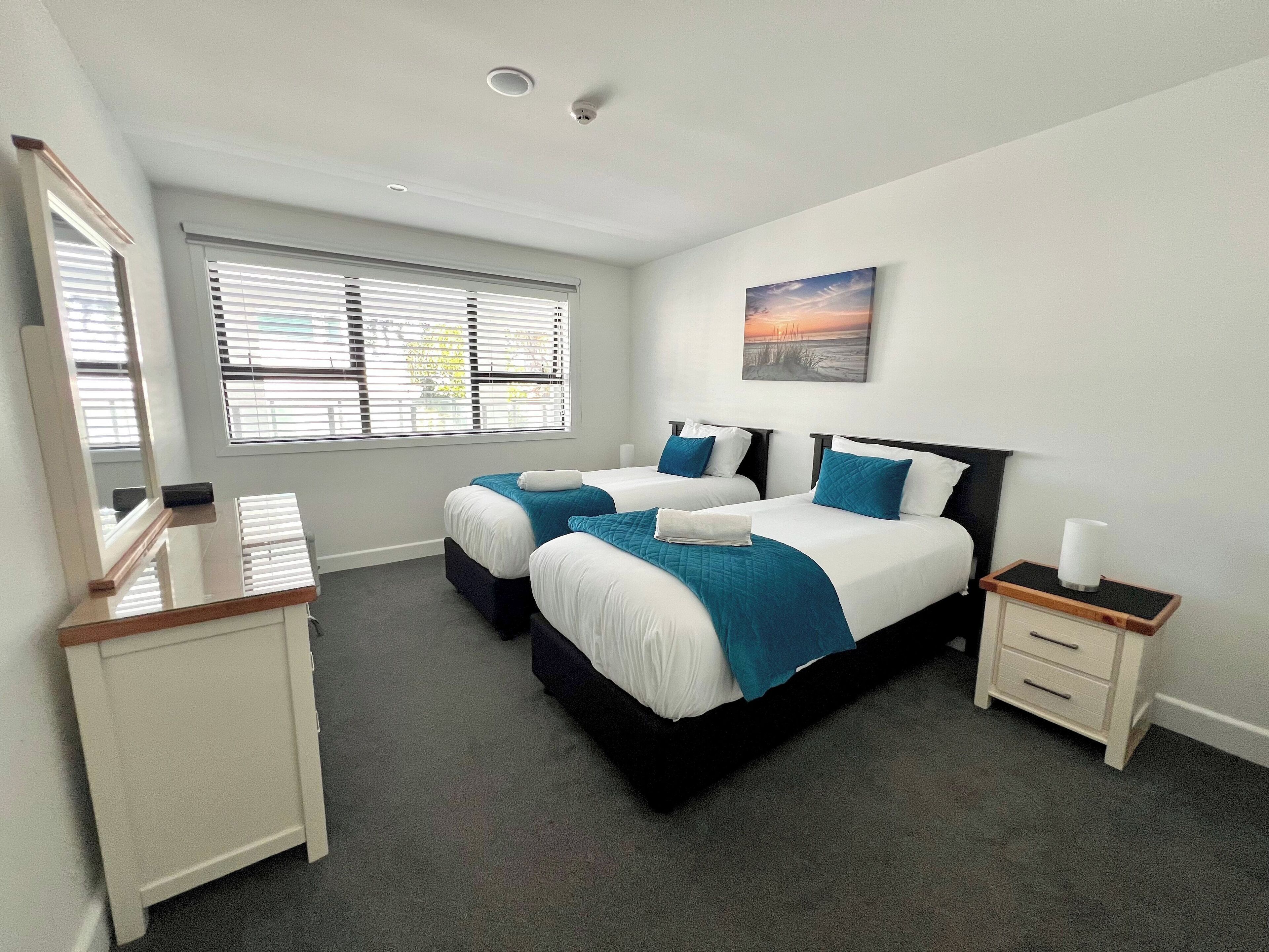 A hotel room with two beds, a desk, a nightstand, and a window with blinds.