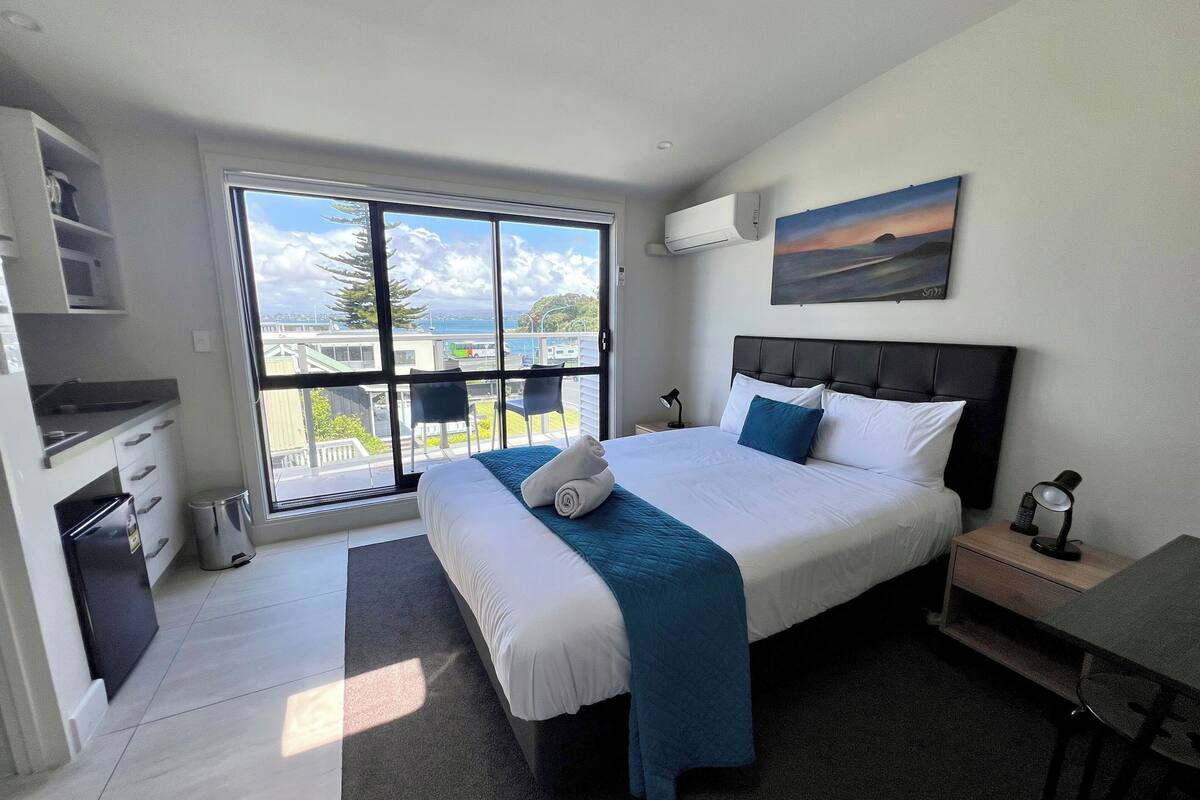 A modern hotel room with a large bed, a desk, a chair, a small table, a nightstand, a lamp, and a painting on the wall.
