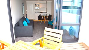 A living area with a sofa, a dining table, and chairs. There is a kitchen in the background. - 6 on Adams  (Tauranga)