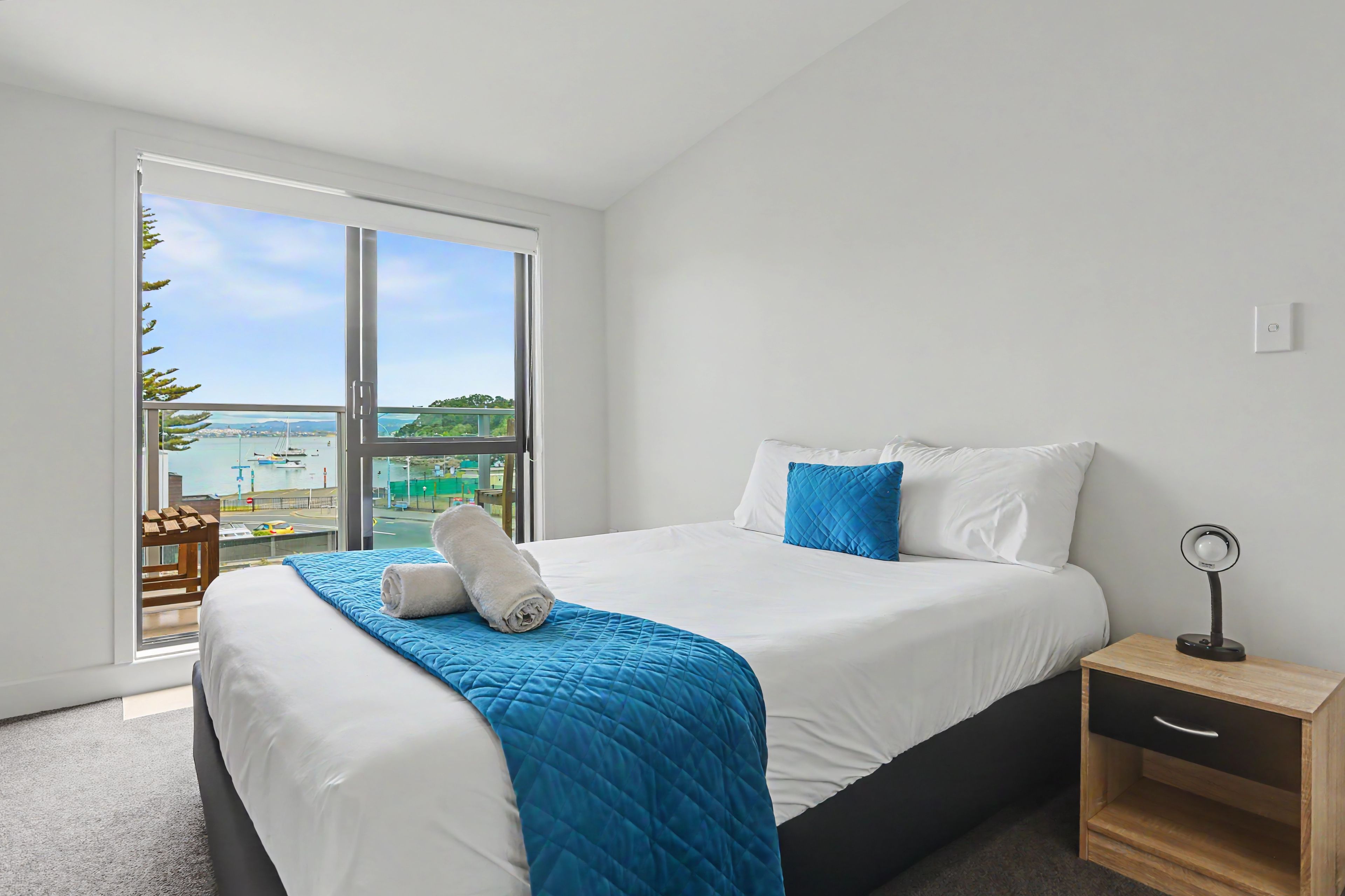 A hotel room with a large bed, a bedside table with a lamp, and a view of the sea through a large window.