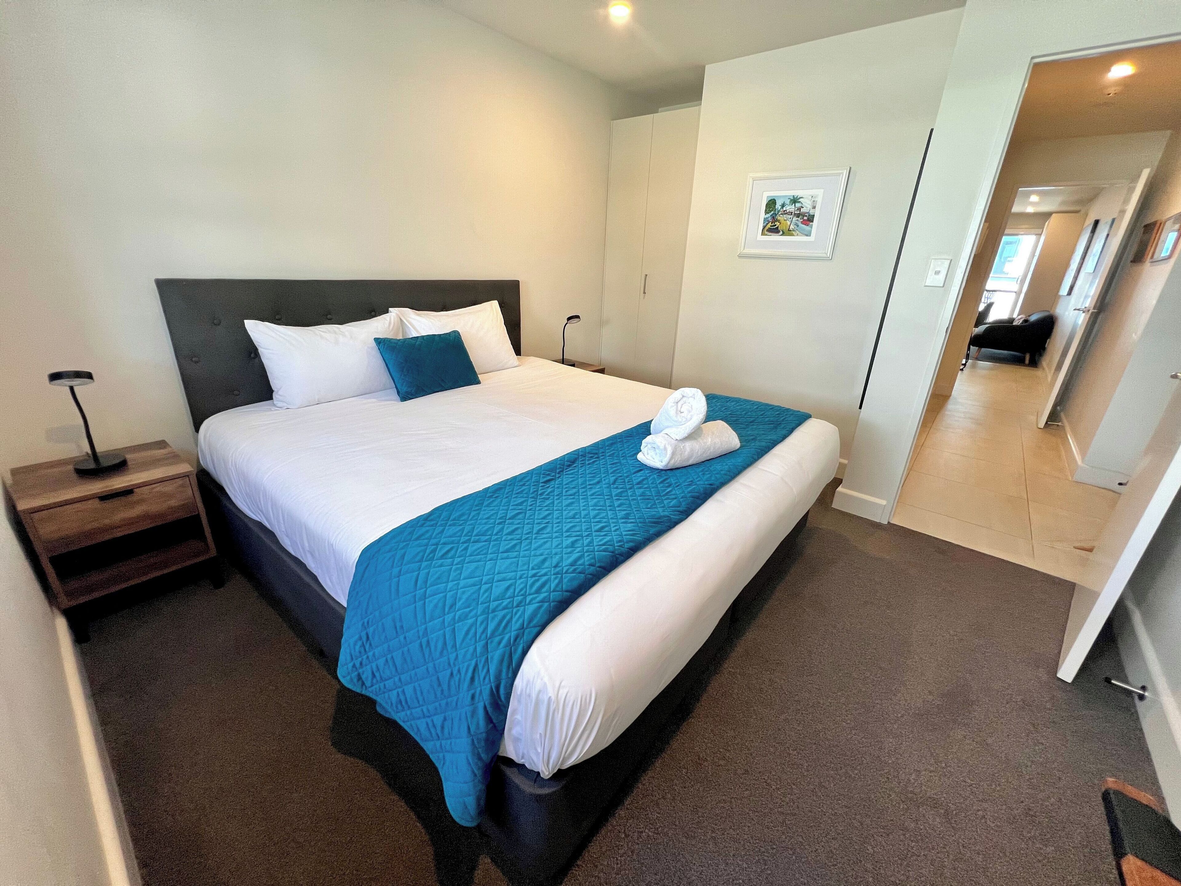 A neatly made bed with a blue quilt, white pillows, and a white towel on the side. There is a small wooden nightstand with a lamp beside the bed.