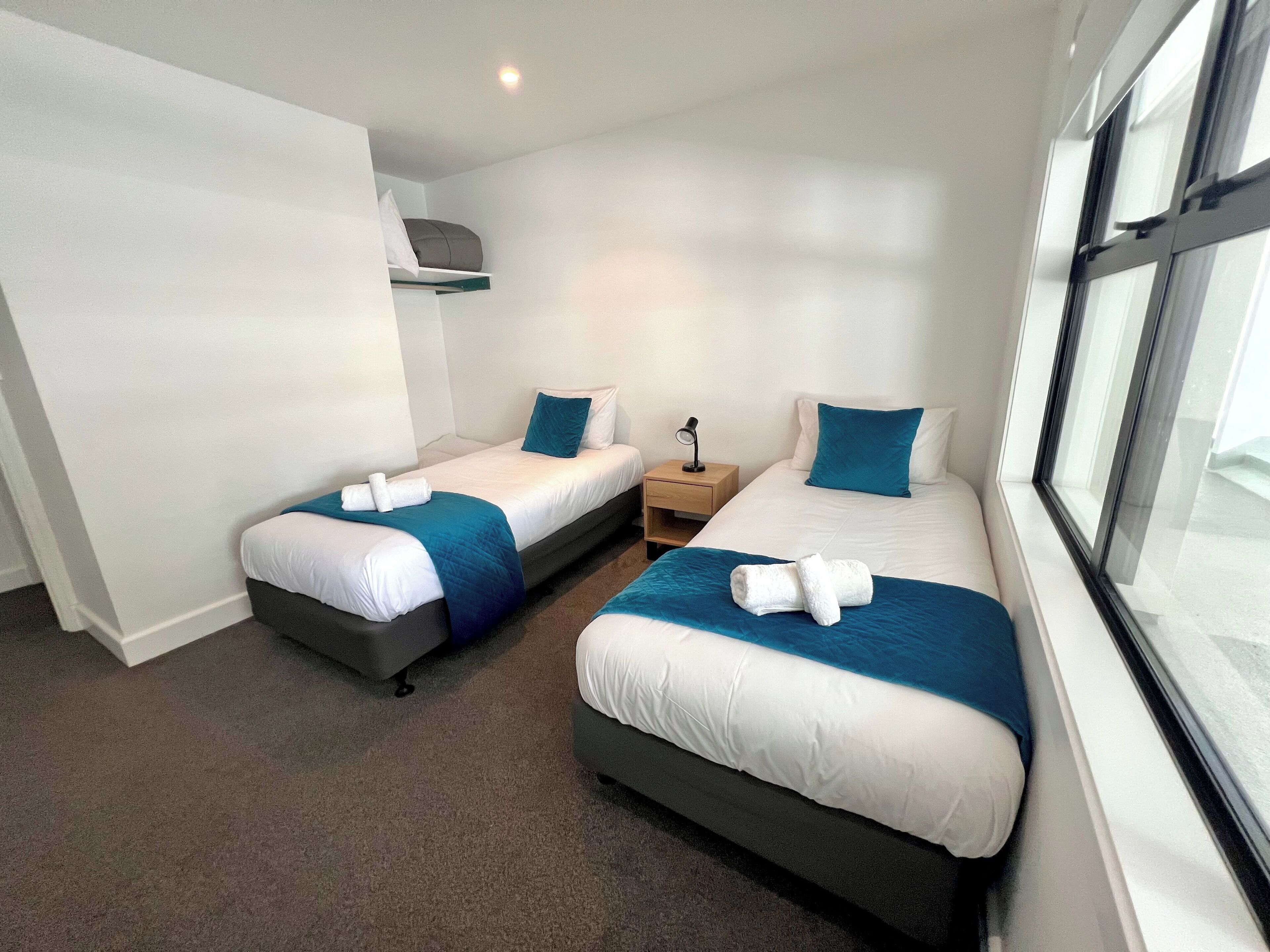 Habitación de hotel con dos camas individuales, cada una con una colcha azul y almohadas blancas, y una mesita de noche pequeña con una lámpara.