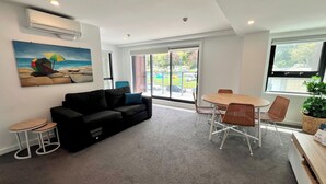 A modern living room with a black sofa, a small round table, and a dining table with chairs. There is a painting on the wall and a potted plant on the right. - 6 on Adams  (Tauranga)