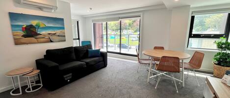 A modern living room with a black sofa, a small round table, and a dining table with chairs. There is a painting on the wall and a potted plant on the right.