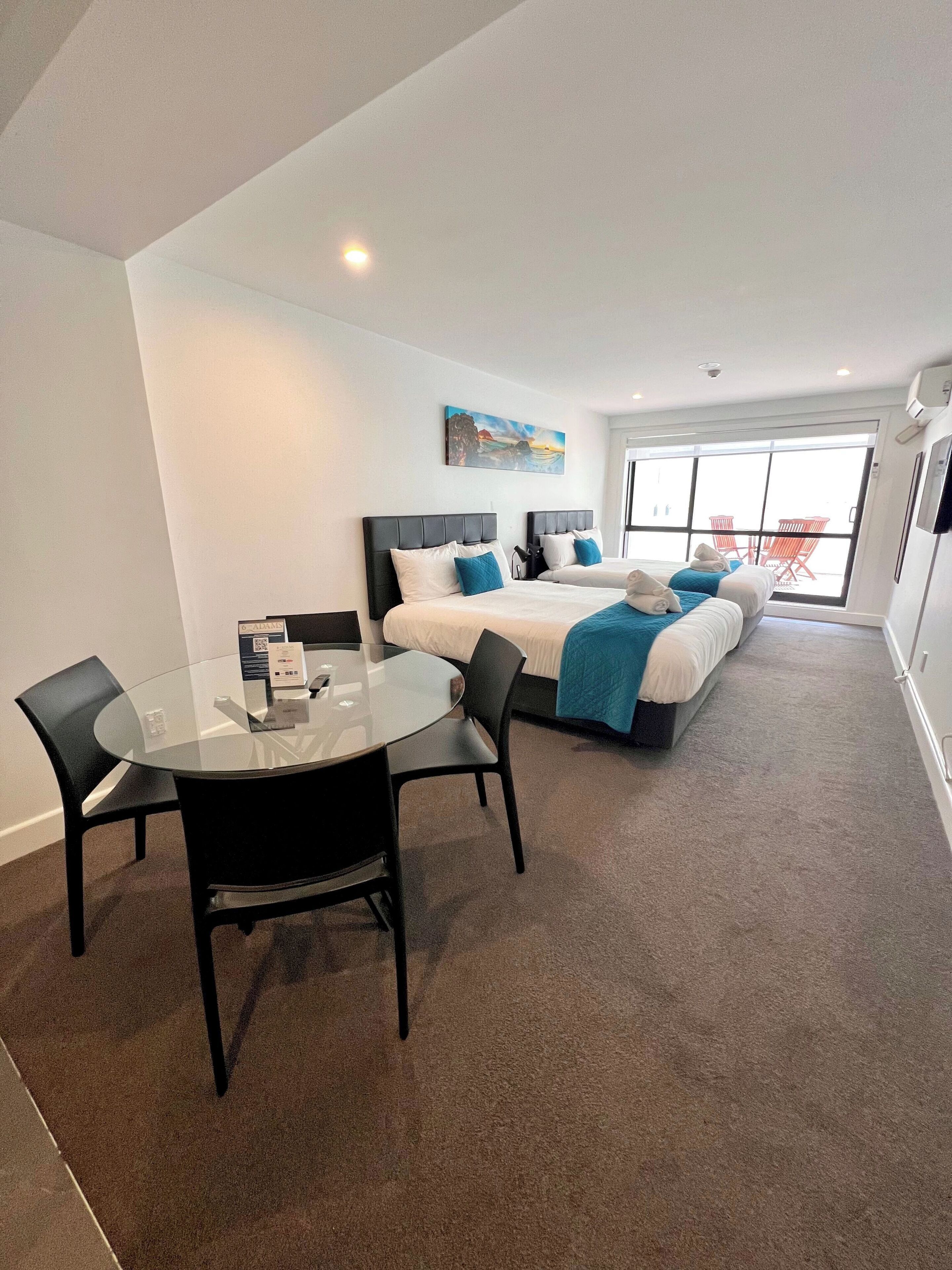 A hotel room with two beds, a glass table, and chairs. There is a painting on the wall and a sliding door leading to an outdoor area.