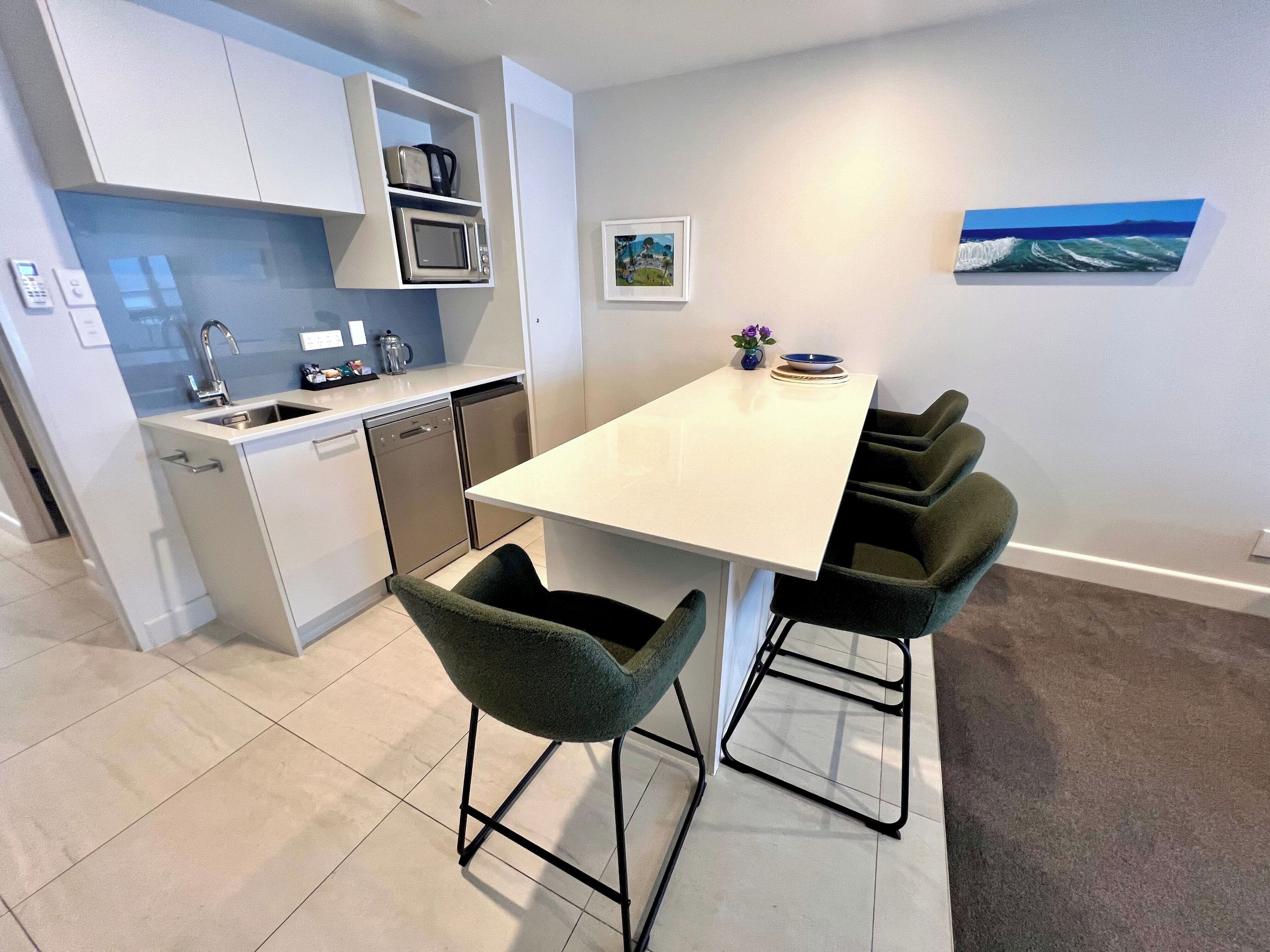 A modern kitchen with white cabinets, a microwave, and a dishwasher. There is a dining area with a white table and four green chairs.