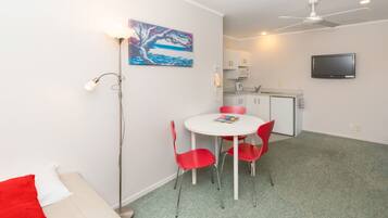 A compact room with a round table, red chairs, a white sofa, a painting, a ceiling fan, a TV, and a kitchenette.