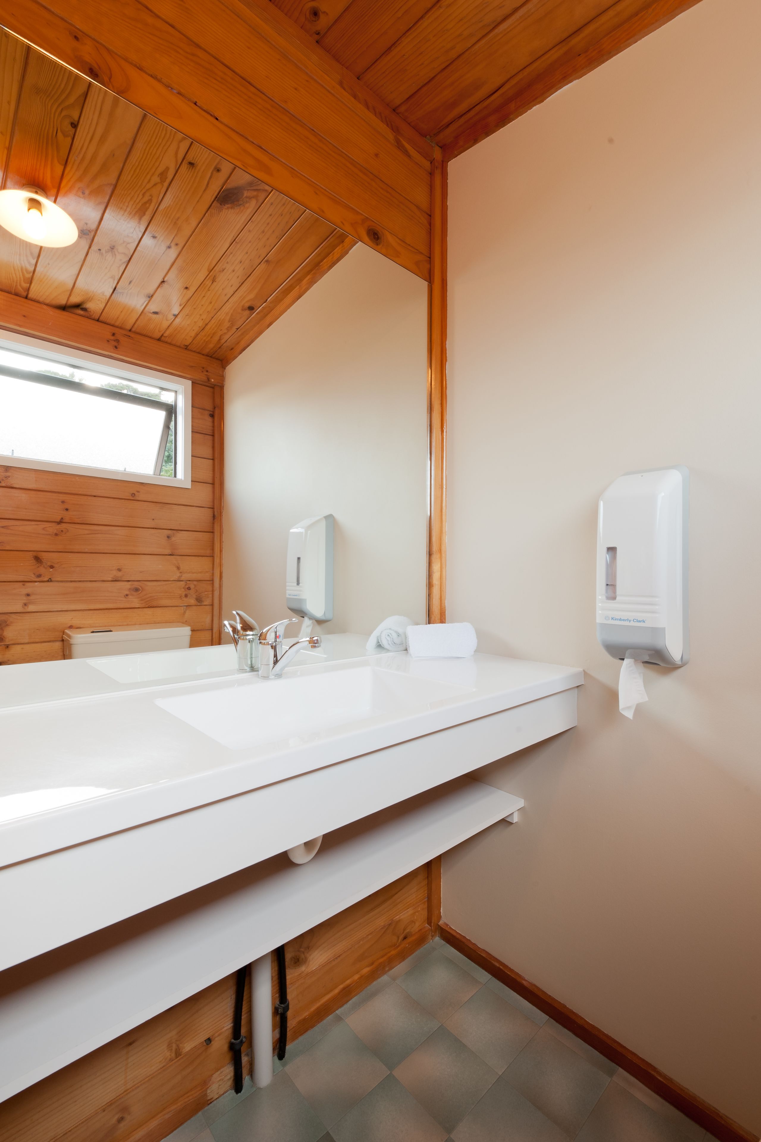 A bathroom with a wooden ceiling, a large white sink, a mirror, and a hand sanitizer dispenser.